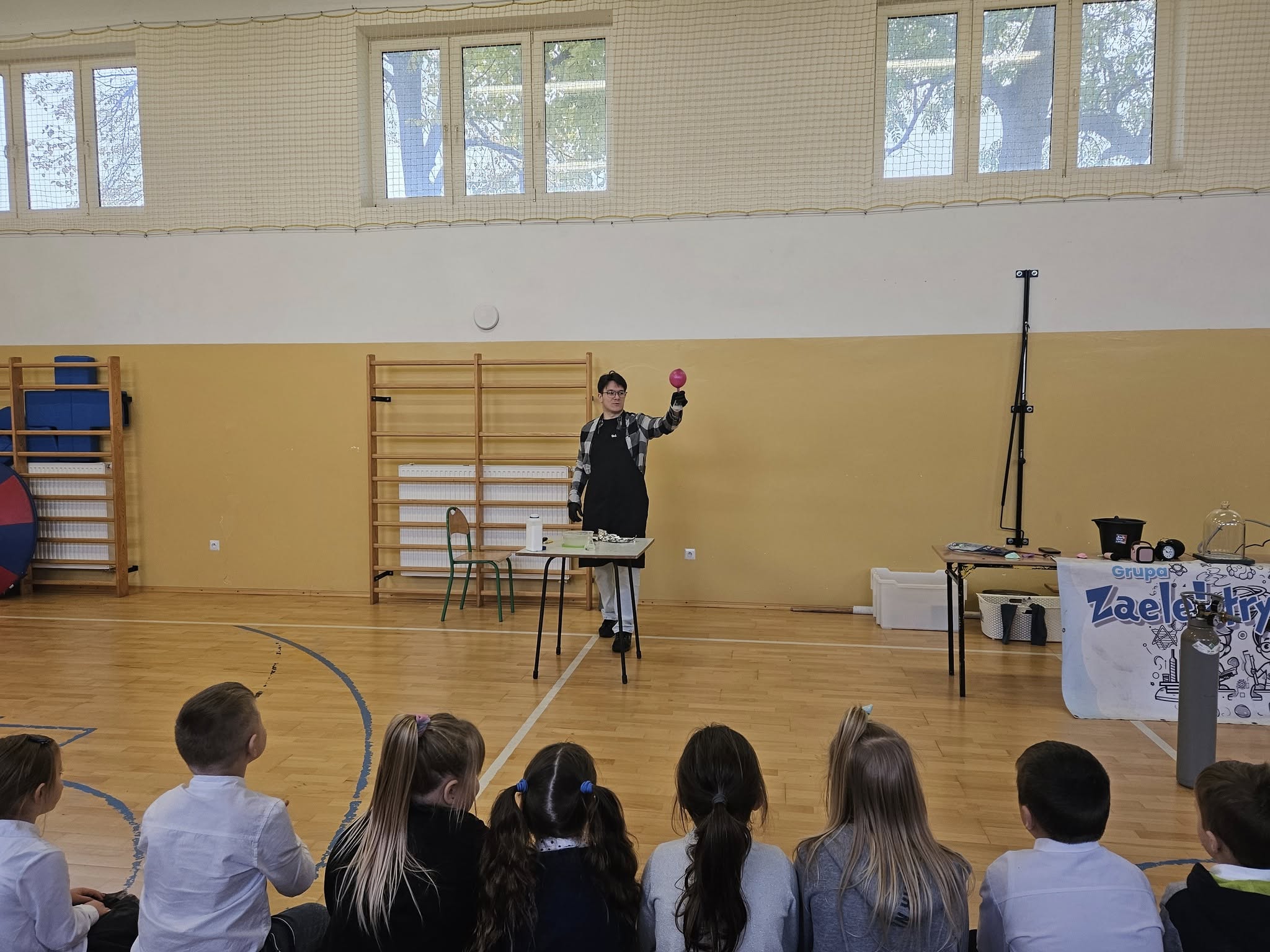 Prowadzący prezentuje eksperyment z fizyki, trzymając różowy balon w uniesionej ręce. Dzieci siedzą naprzeciwko, z zainteresowaniem obserwując przebieg doświadczenia.