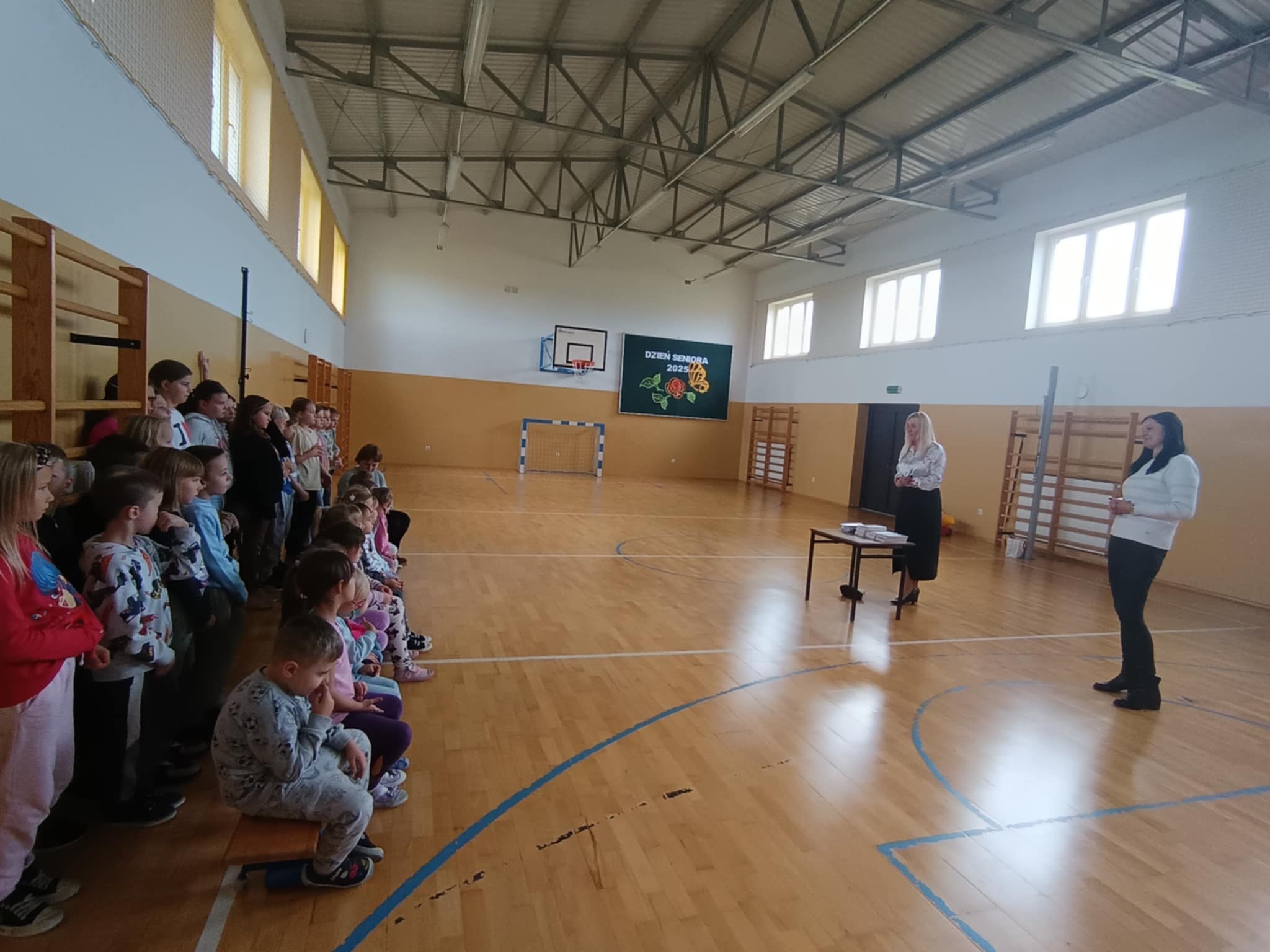 Uczniowie i dzieci z oddziału przedszkolnego uczestniczą w spotkaniu z przedstawicielką Banku Pekao S.A. na sali gimnastycznej.