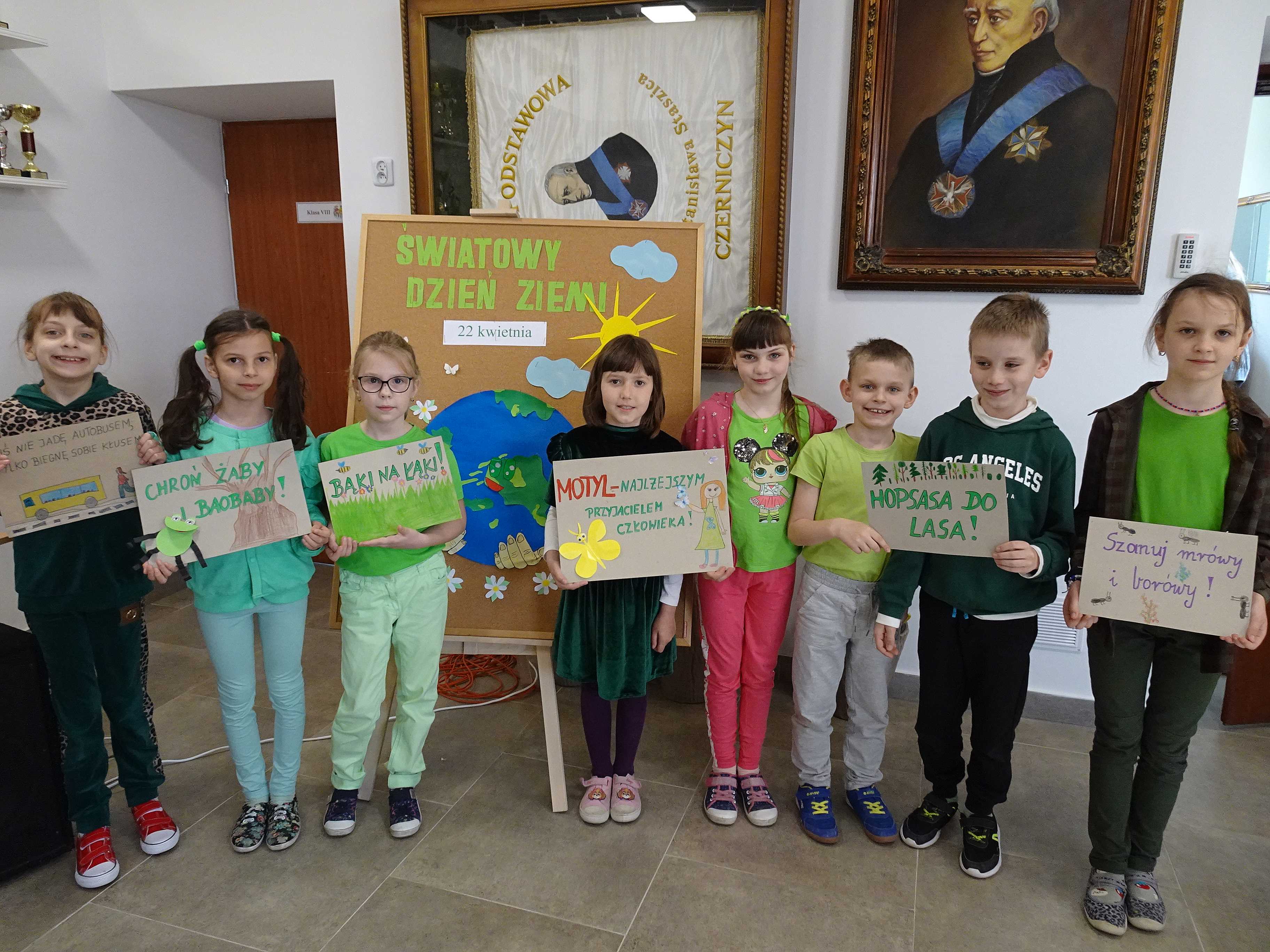 Uczniowie klasy drugiej z plakatami promującymi ochronę środowiska na tle tablicy okolicznościowej z okazji Dnia Ziemi