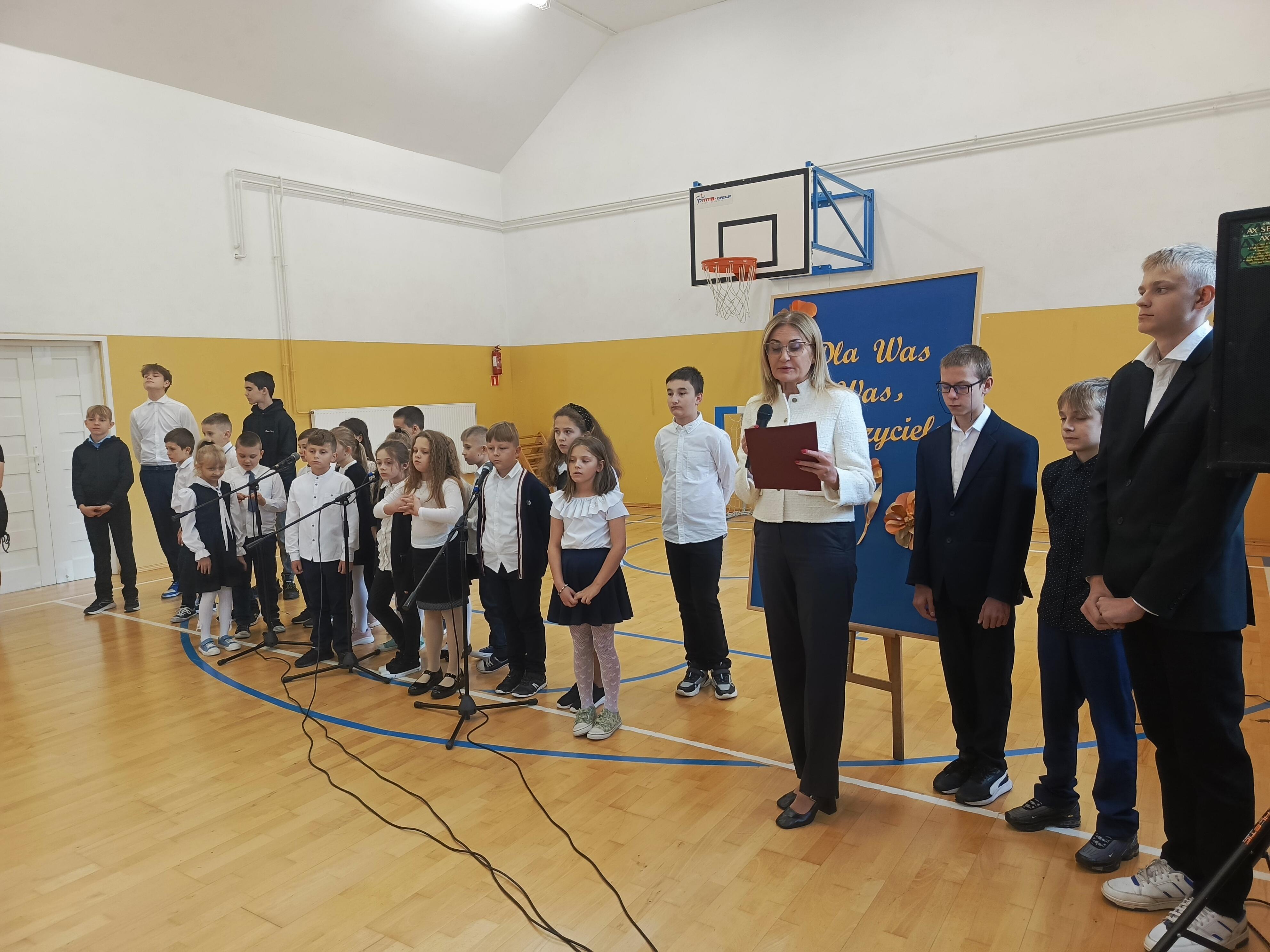 Pani Dyrektor przemawia stojąc przed grupą uczniów w strojach galowych