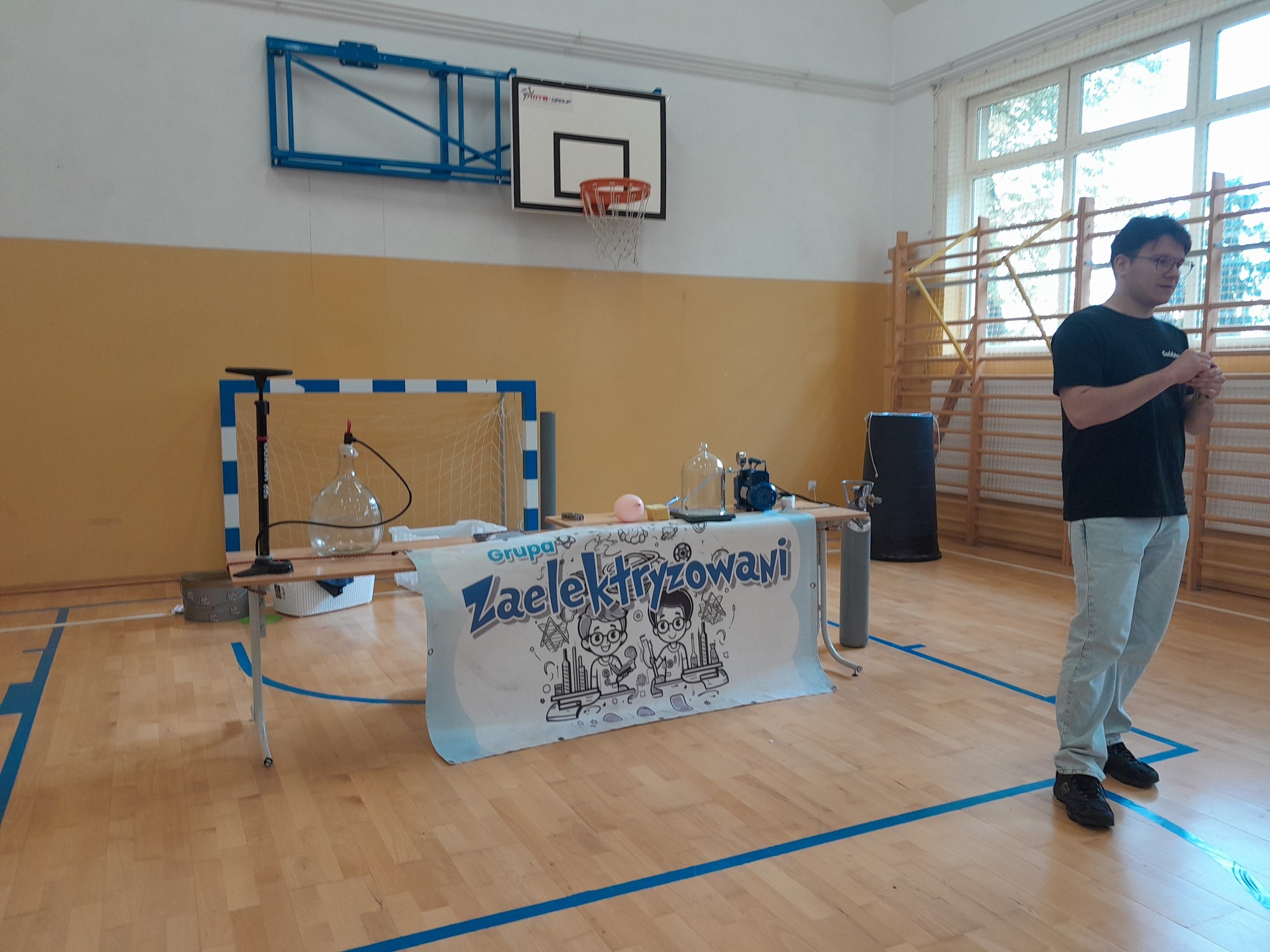 Przedstawiciel grupy &quot;Zaelektryzowani&quot; przy mikrofonie, za nim stolik z przyborami potrzebnymi do eksperymentów