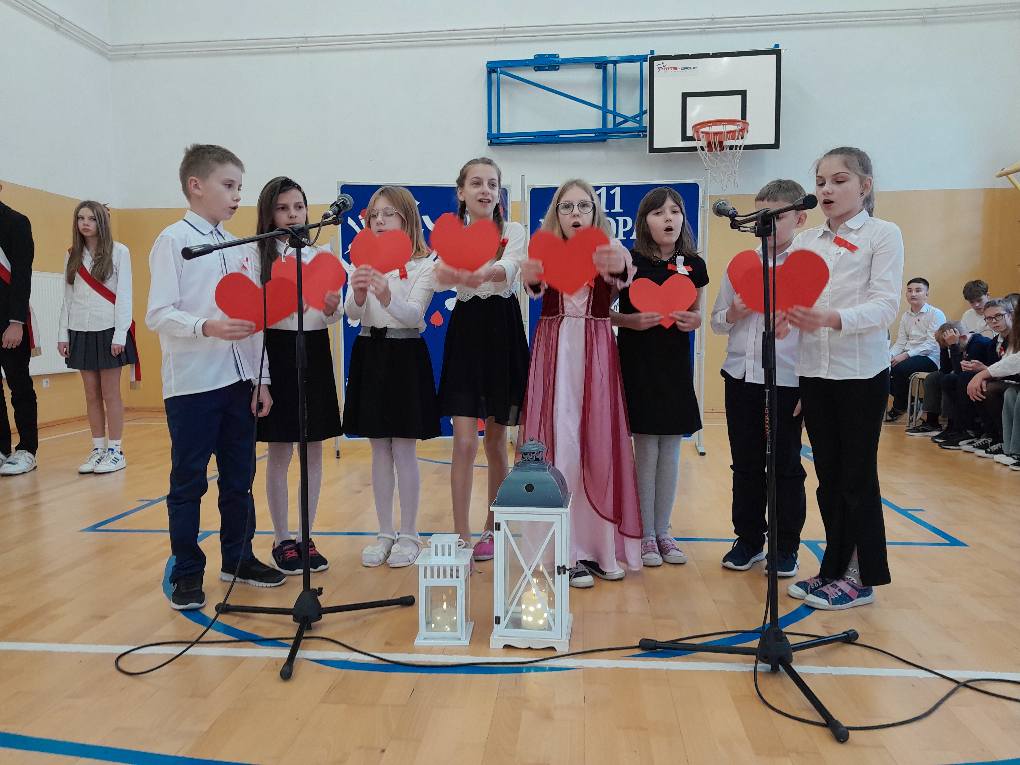 Uczniowie przy mikrofonach, trzymają czerwone papierowe serca w dłoniach