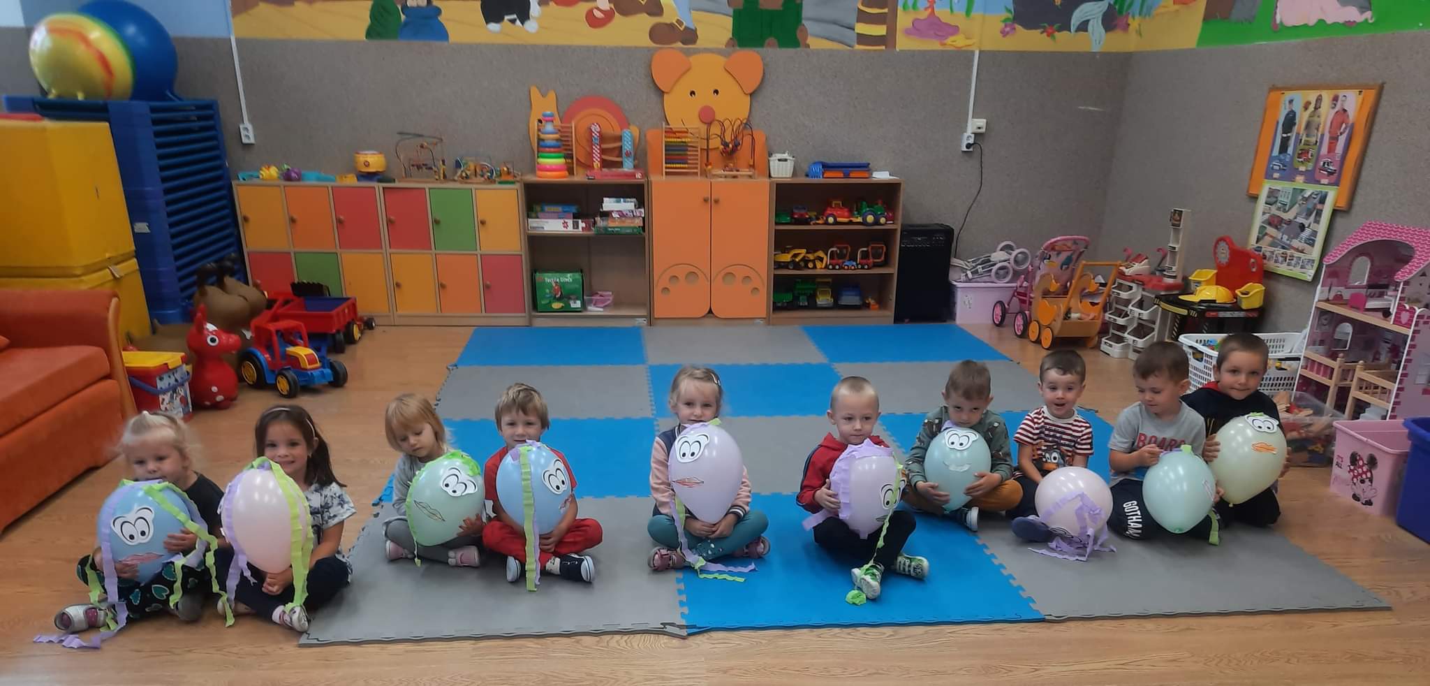 Przedszkolaki - chłopcy i dziewczynki siedzą na dywanie i trzymają kolorowe baloniki w rękach