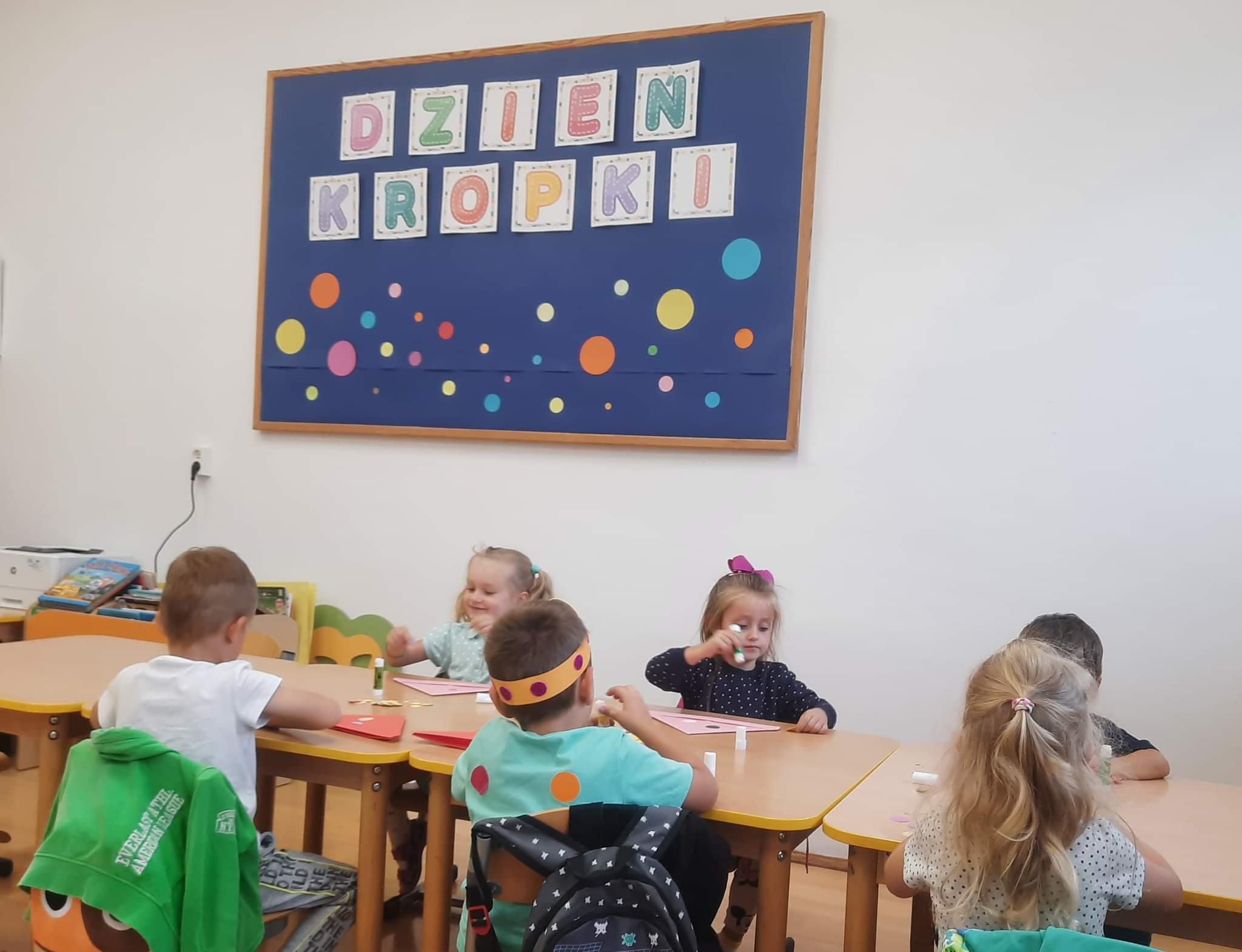 Chłopcy i dziewczynki - przedszkolaki przy stolikach ozdabiają papierowe czapeczki kolorowymi kropkami
