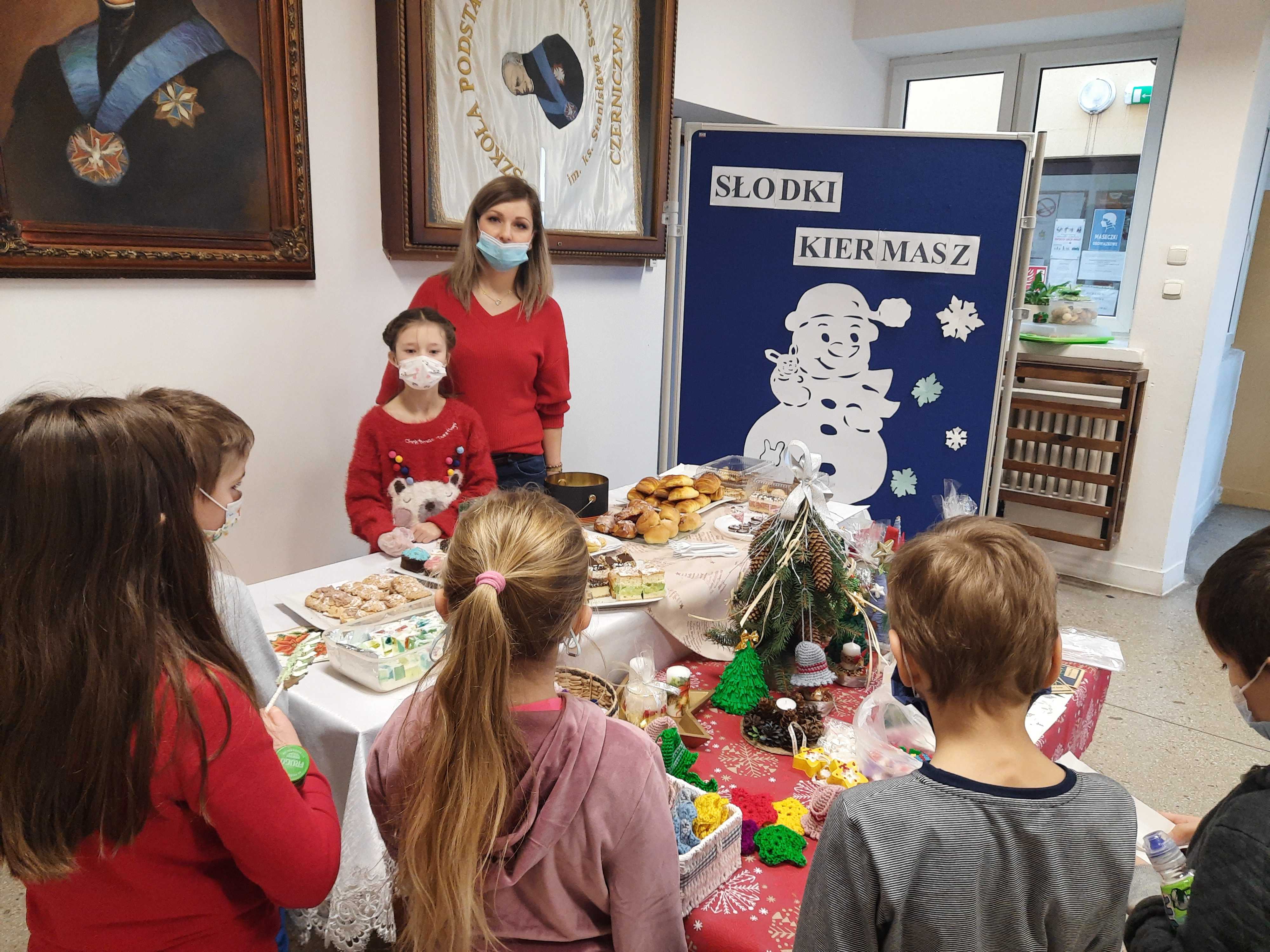 Nauczycielka i uczennica za stolem z ciastami i ozdobami świątecznymi. Uczniowie zgromadzeni dookoła stołu