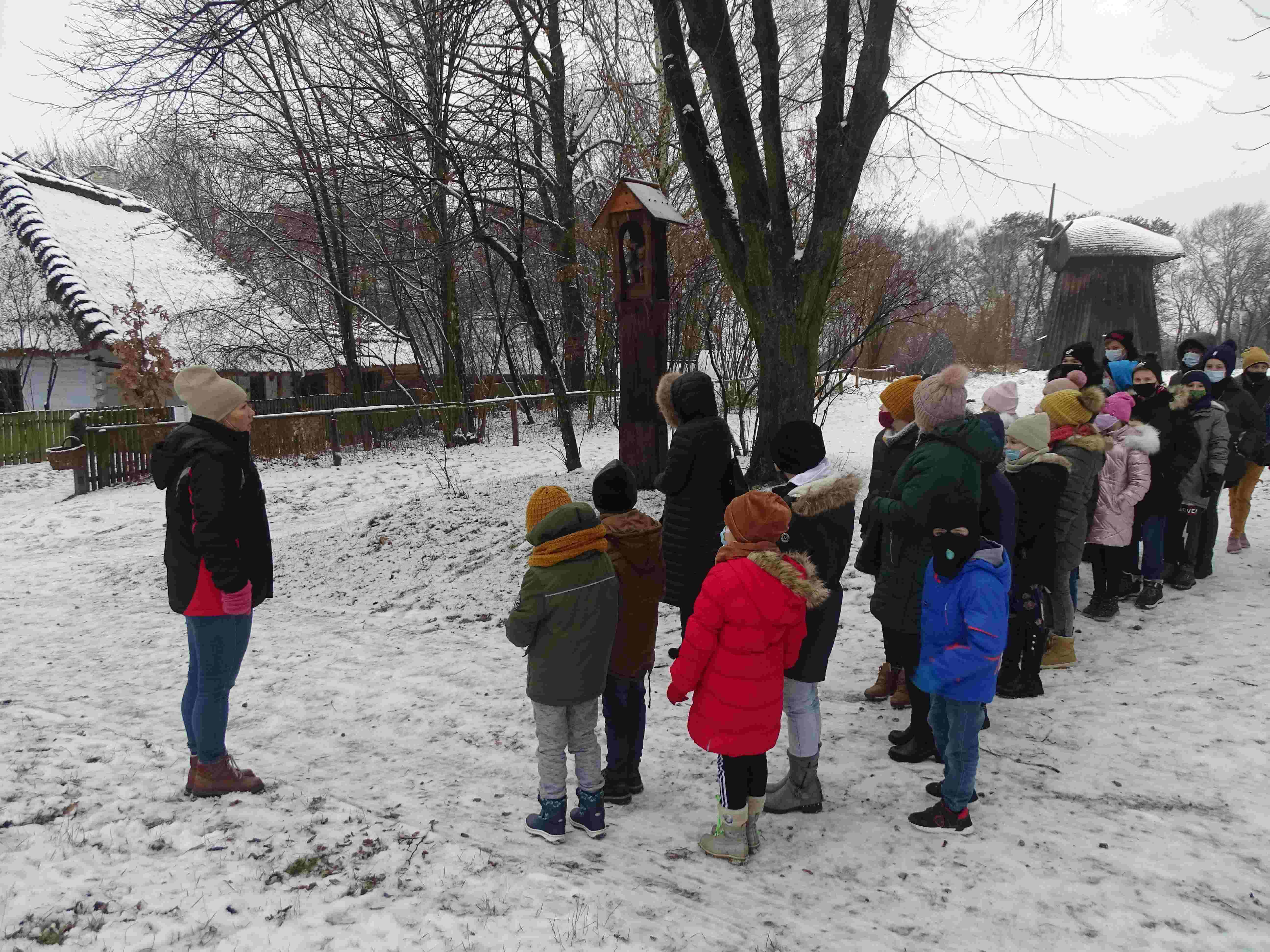 Przewodniczka z grupą uczniów i nauczycielkami przy wiejskiej drewnianej kapliczce. W tle wiejskie chaty