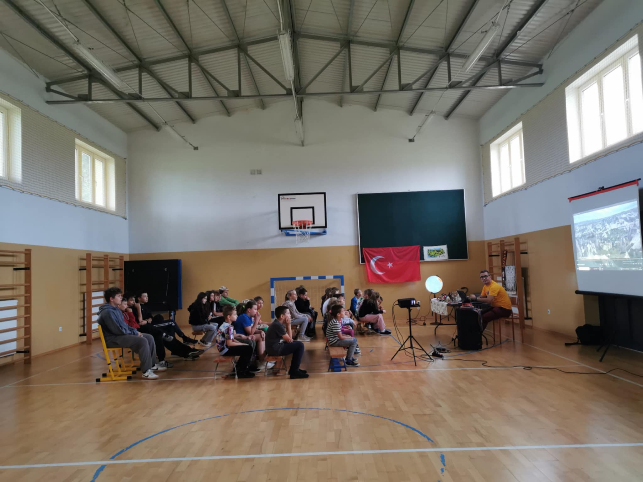  Ujęcie całej sali gimnastycznej – uczniowie siedzą na ławkach, oglądają prezentację wyświetlaną na ekranie.