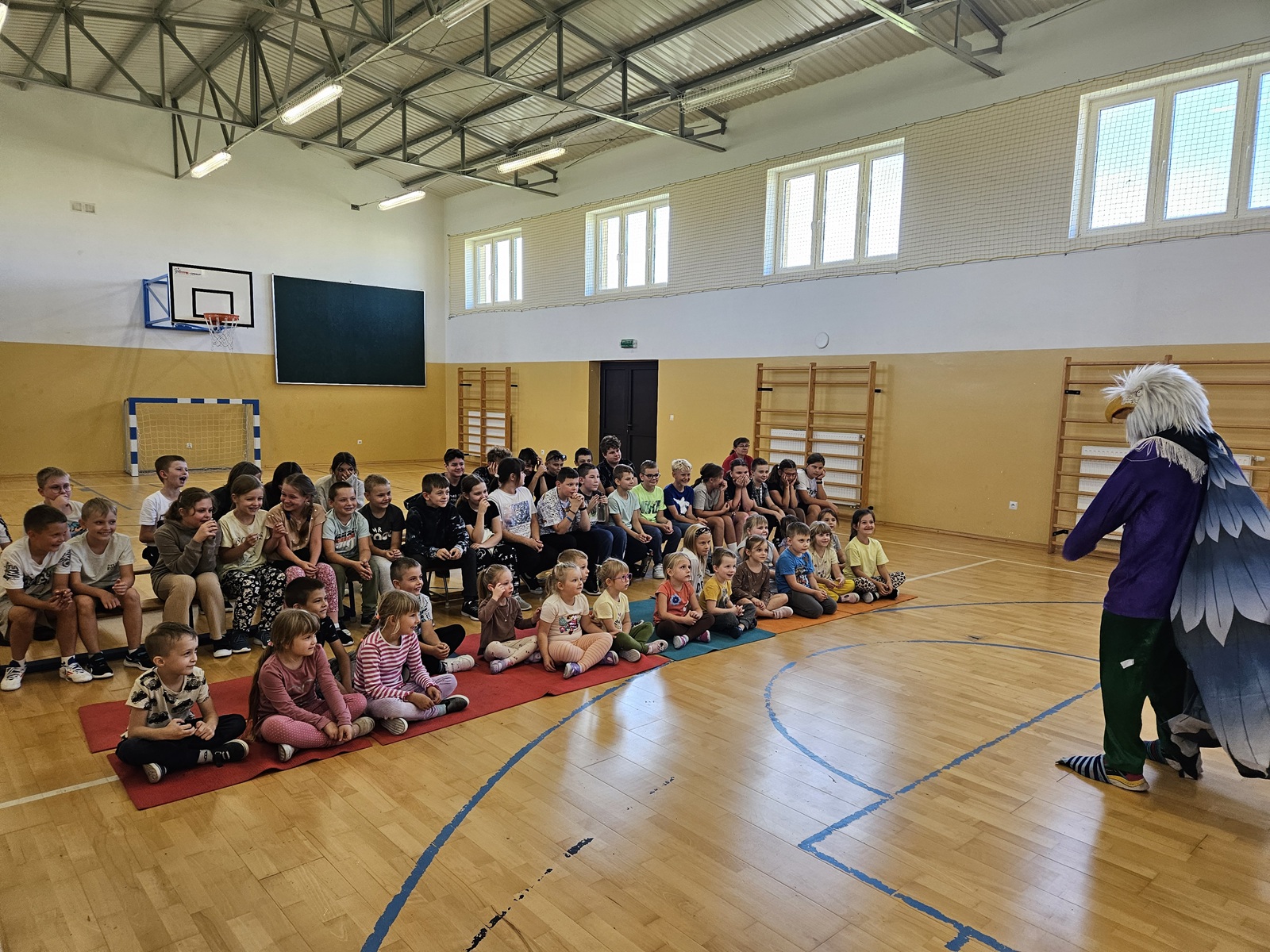 Klaun Walduś w kostiumie z dużymi skrzydłami prezentuje pokaz przed dziećmi na sali gimnastycznej.
