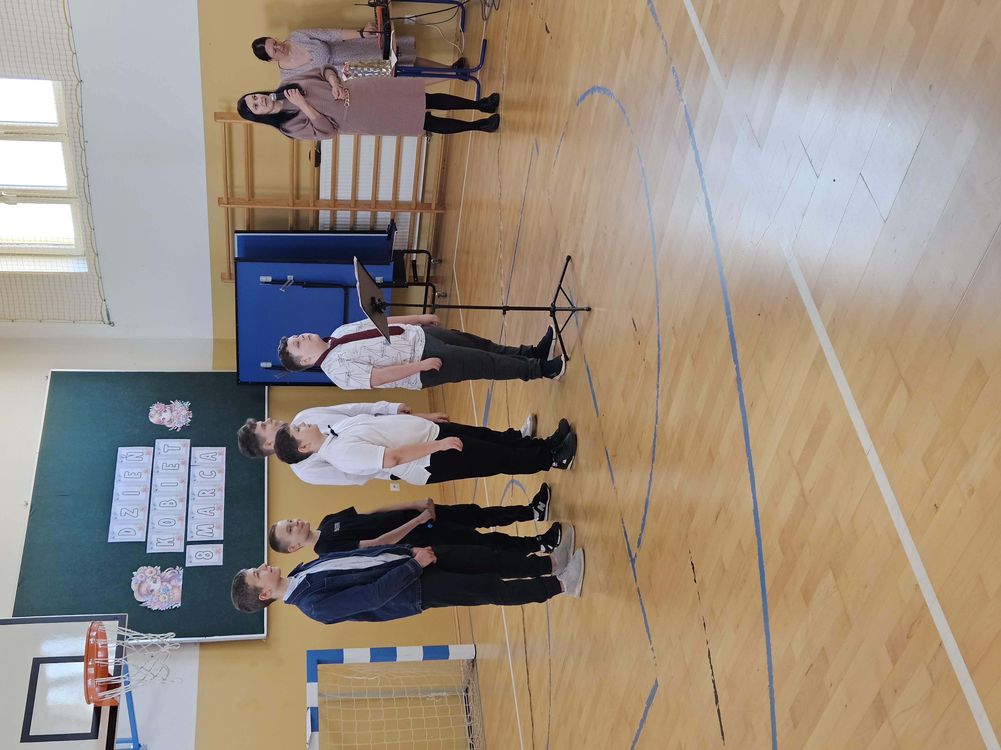 Chłopcy stoją na środku sali gimnastycznej przy mikrofonie podczas występu z okazji Dnia Kobiet. W tle widoczna jest dekoracja z napisem „Dzień Kobiet – 8 marca”, a obok stoi pani dyrektor oraz pani prowadząca uroczystość.