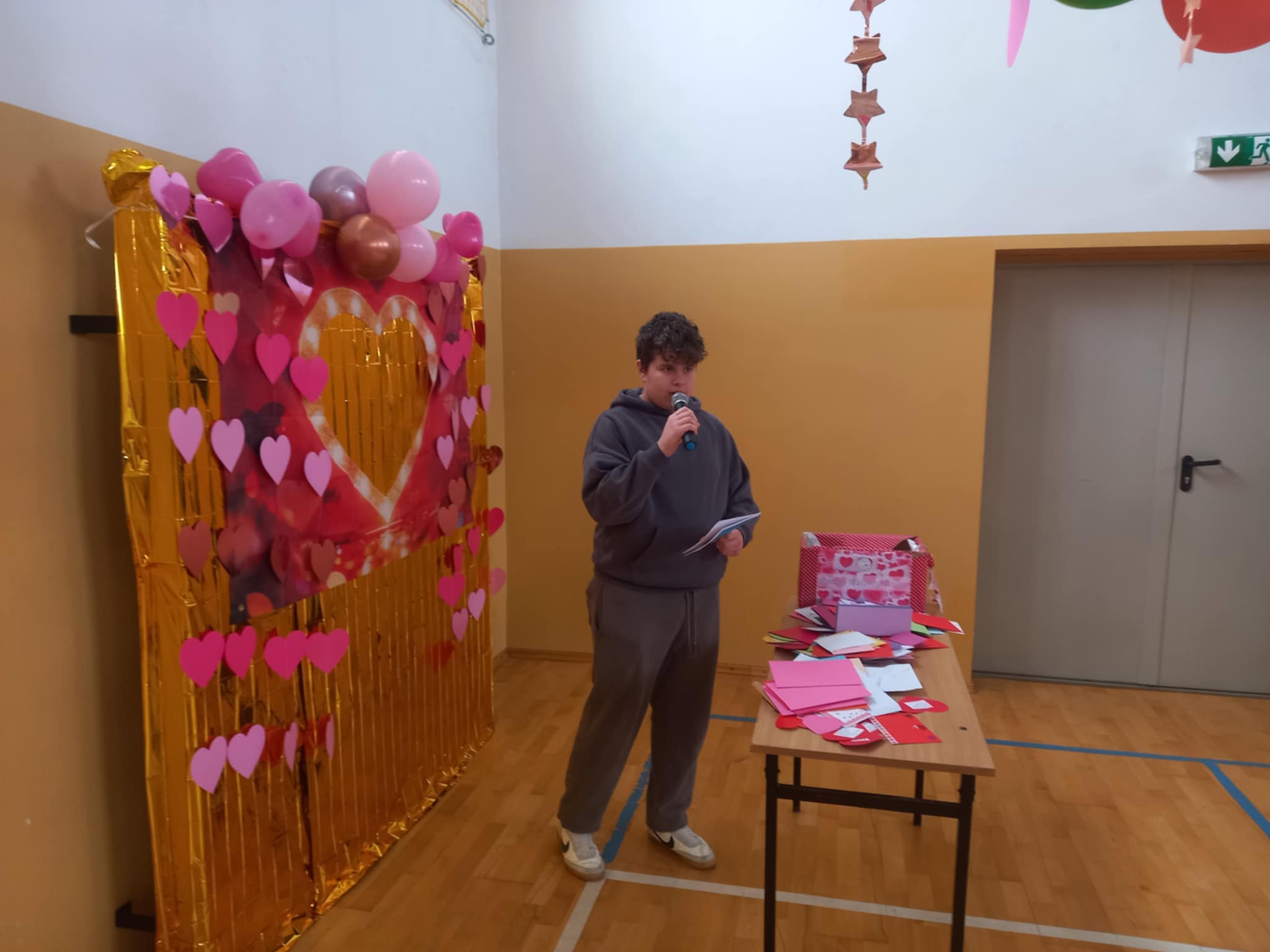 Uczeń z mikrofonem stoi przy stoliku z kartkami walentynkowymi; obok znajduje się dekoracyjna ścianka z motywem serca i balonami.