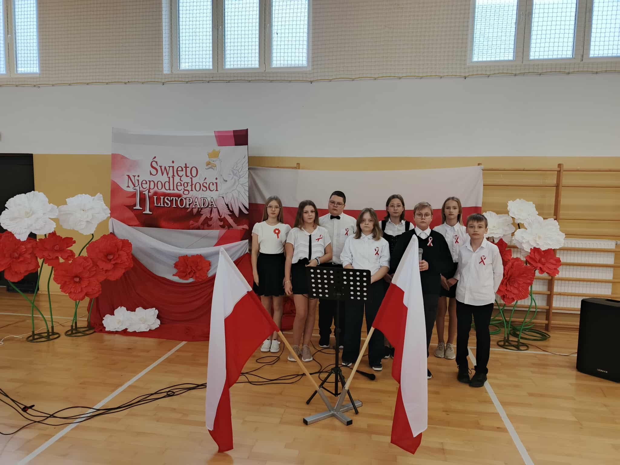 Uczniowie i nauczyciele stoją w sali gimnastycznej podczas akademii z okazji Święta Niepodległości.