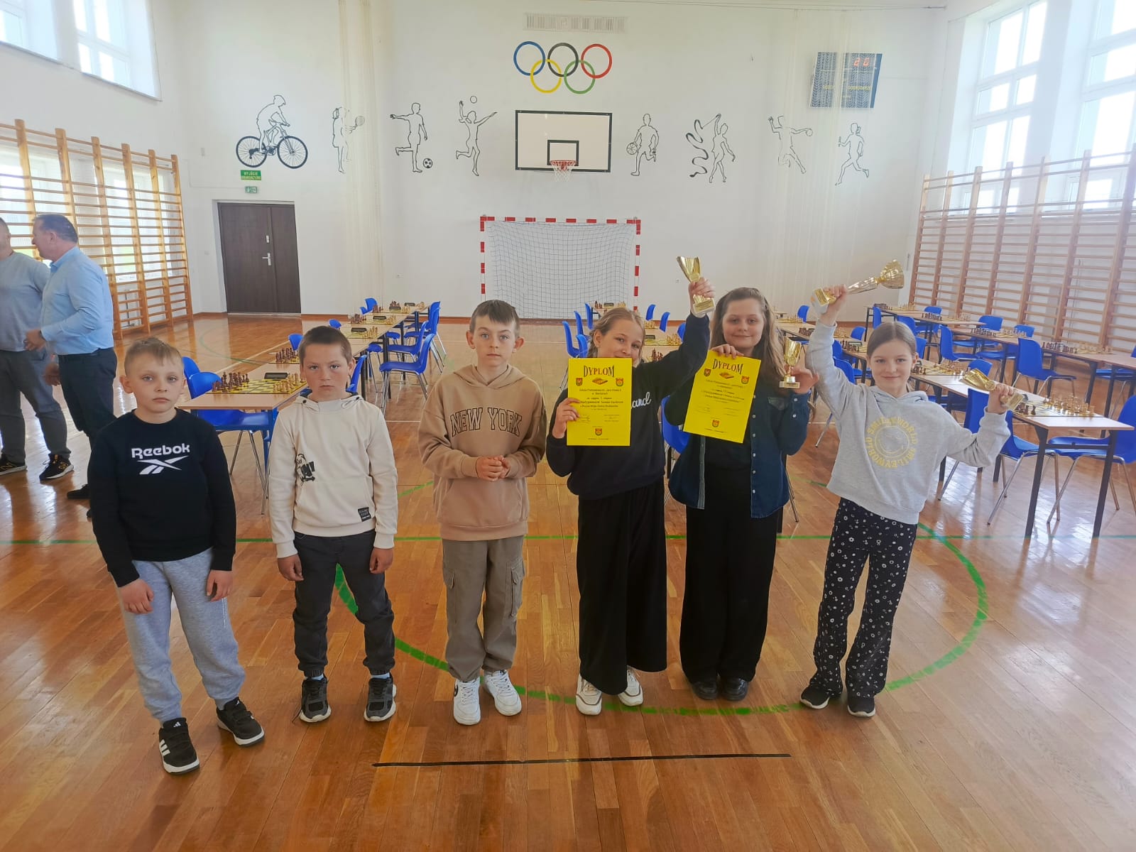 Reprezentacja Szkoły Podstawowej w Mienianach po zakończeniu turnieju szachowego pozuje z dyplomami i pucharami na sali gimnastycznej.