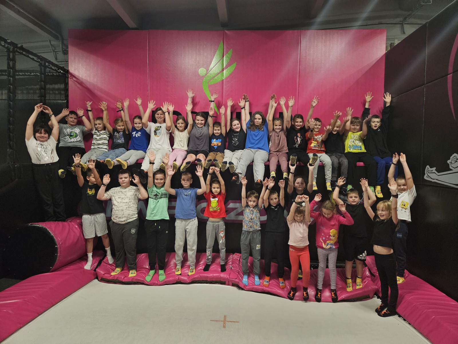 Grupa uczniów pozująca wspólnie na konstrukcji w parku trampolin; dzieci siedzą i stoją na tle różowej ściany.
