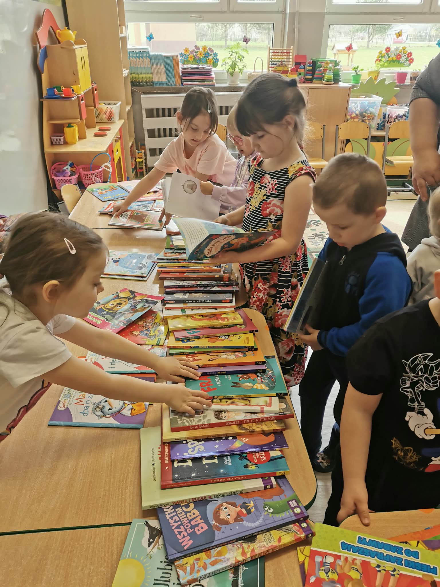 Dzieci oglądają książki rozłożone na stoliku, wybierając pozycje do wypożyczenia.