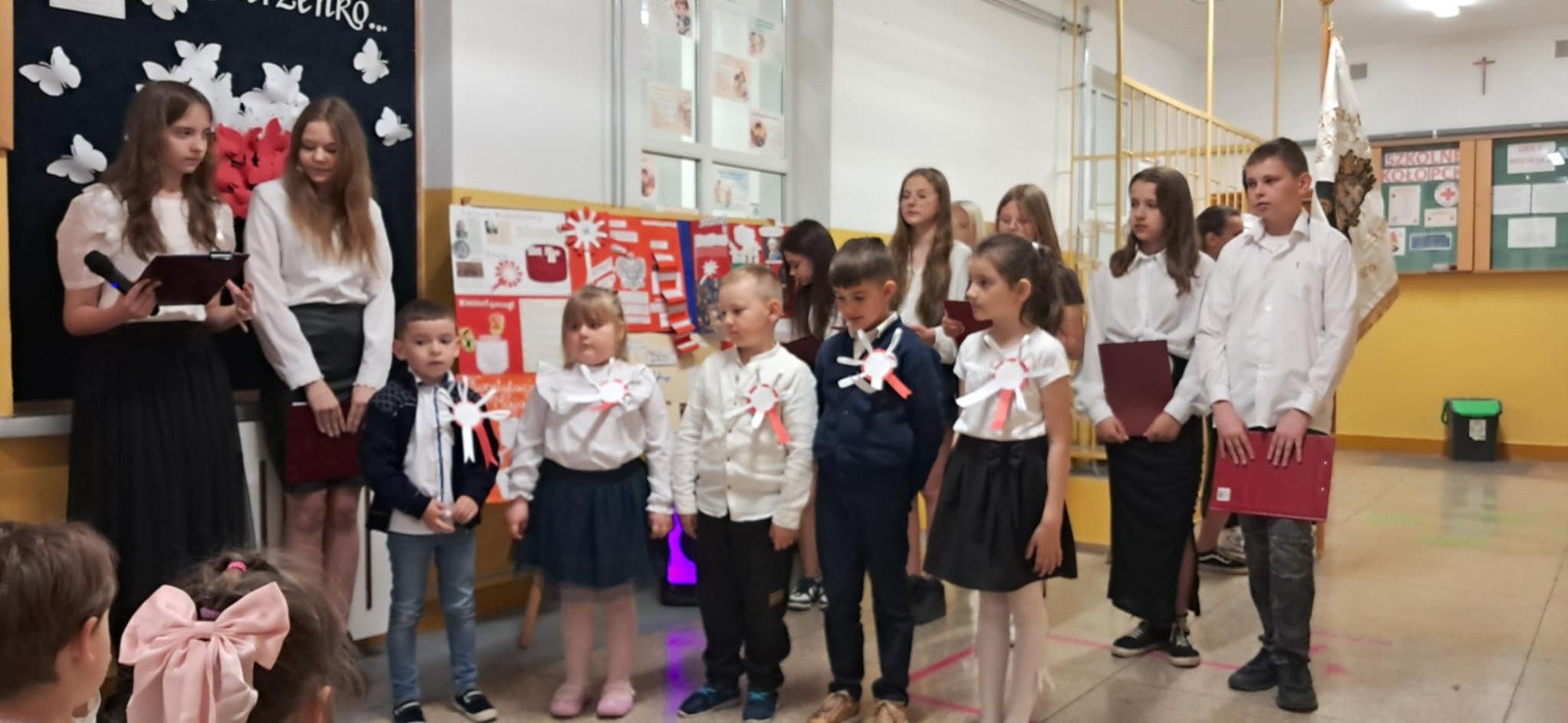 Uczniowie szkoły podstawowej wraz z dziećmi z oddziału przedszkolnego biorą udział w akademii z okazji świąt majowych.