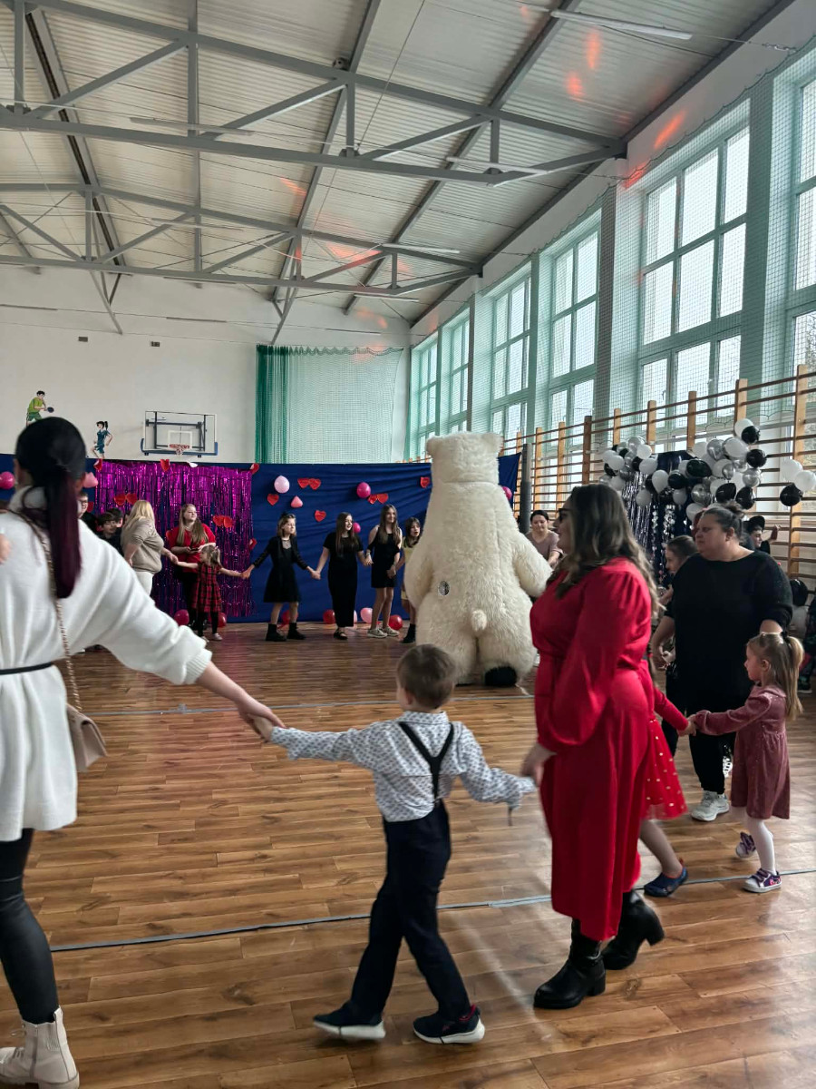 Dzieci wraz z uczniami biorą udział w zabawie choinkowej przy muzyce, a w środku tańczy biały miś.