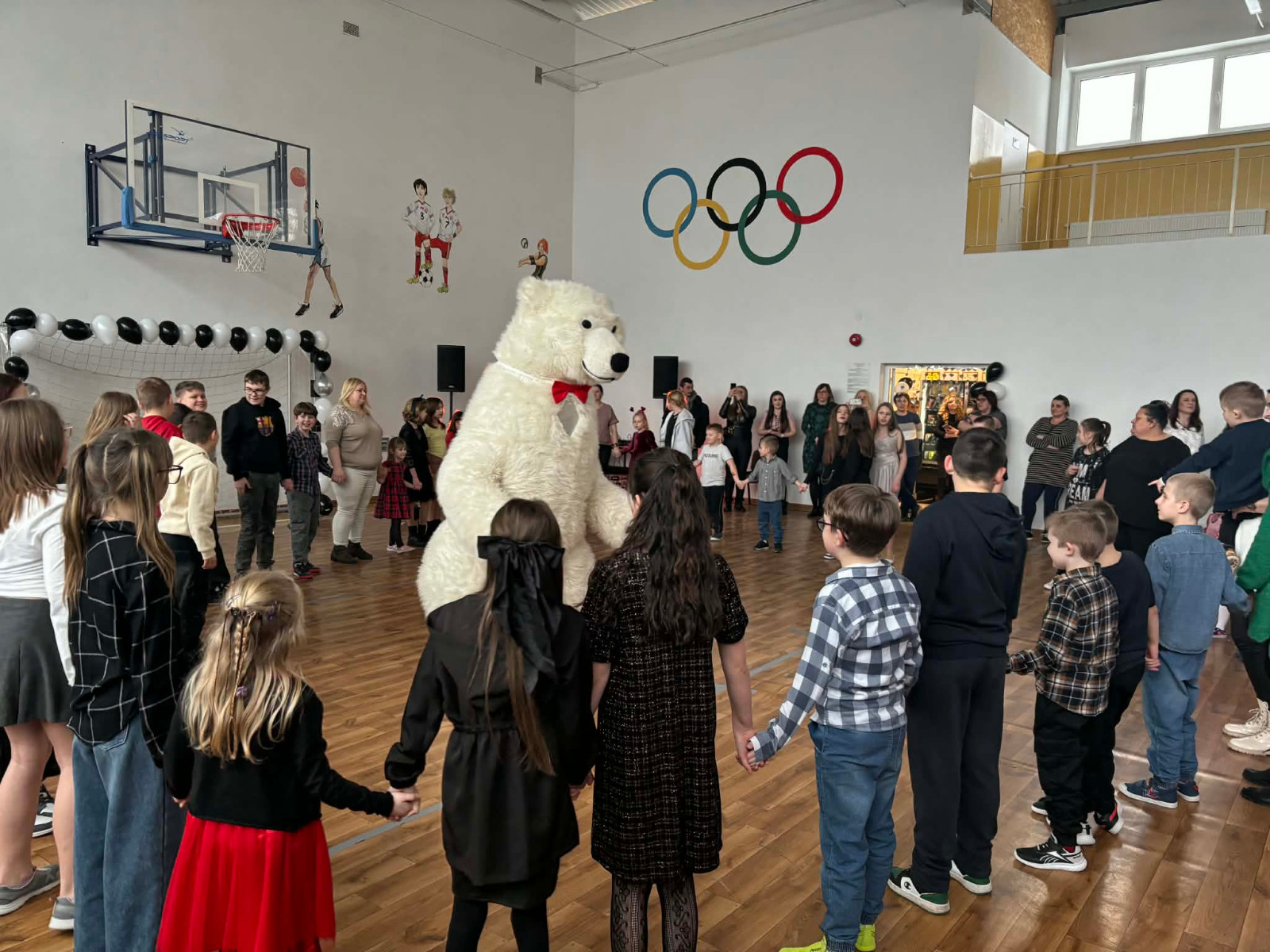 Dzieci wraz z uczniami biorą udział w zabawie choinkowej przy muzyce, a w środku tańczy biały miś.
