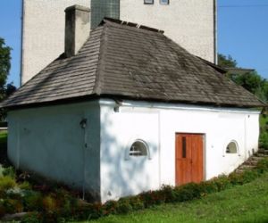 Biały budynek z półokrągłymi okienkami, drewnianymi drzwiami i czarnym dachem pokrytym dachówką 