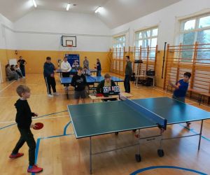 Dwóch chłopców rozgrywa mecz tenisa stołowego, a trzeci sędziuje. W tle trwają mecze przy dwóch kolejnych stołach
