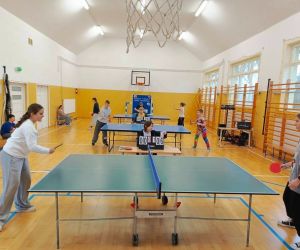 Dwie dziewczynki rozgrywają mecz tenisa stołowego, a chłopiec sędziuje. W tle trwają mecze przy dwóch kolejnych stołach