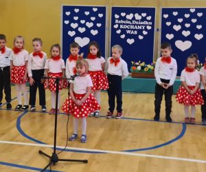 Dziewczynka recytuje przy mikrofonie, za nią stoją w szeregu chłopcy i dziewczynki