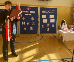 Uczeń stoi przy mikrofonie, a w tle dziewczynka siedzi przy stoliku