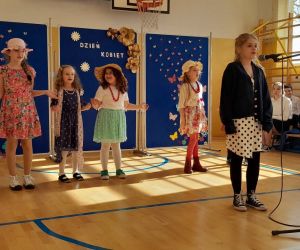 Uczennica klasy czwartej śpiewa do mikrofonu, za nią tańczą dziewczynki z przedszkola w kolorowych strojach