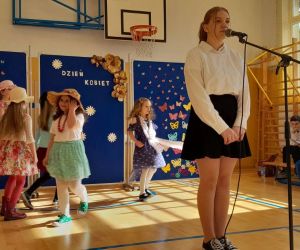 Uczennica klasy ósmej śpiewa do mikrofonu, za nią tańczą dziewczynki z przedszkola w kolorowych strojach