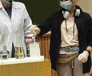 Uczennica klasy ósmej i nauczycielka za pulpitem. Uczennica miesza pipetą substancję chemiczną w szklanym naczyniu