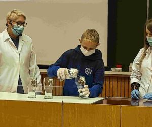 Uczeń klasy ósmej i dwie nauczycielki za pulpitem. Uczeń w rękawiczkach przelewa substancję chemiczną z jednego szklanego naczynia do drugiego