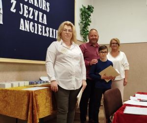 Uczeń z dyplomem w dłoniach, pani Dyrektor szkoły w Husynnem, nauczycielka i pan Inspektor Oświaty na tle tablicy dekoracyjnej