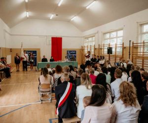 Dyrektor szkoły stoi przy mikrofonie, po jej prawej stronie Poczet Sztandarowy, a przed nią uczniowie siedzą na ławkach