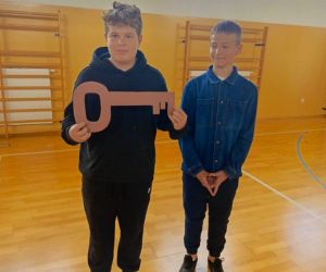 Dwóch chłopców stoi na parkiecie sali gimnastycznej. Jeden z nich trzyma symboliczny klucz, drugi stoi obok i patrzy na kolegę.