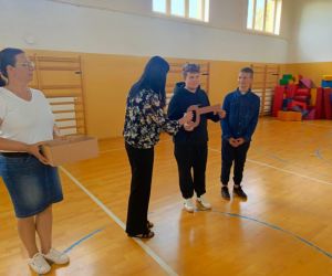 Dwóch uczniów stoi na sali gimnastycznej. Jeden z nich trzyma w rękach duży, symboliczny kartonowy klucz. Obok nich stoją dwie nauczycielki, jedna trzyma pudełko.