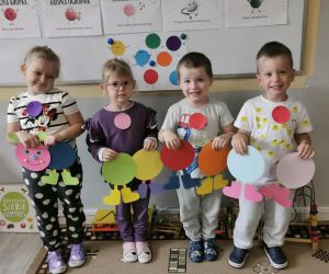 Czworo dzieci stoi pod tablicą z pracami i kolorowymi dekoracjami w kształcie kropek. Dzieci uśmiechają się i pozują do zdjęcia.