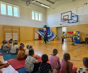 Klaun Walduś w stroju z dużymi skrzydłami występuje przed całą grupą dzieci i uczniów siedzących na sali gimnastycznej.