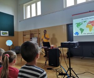 Podróżnik Mateusz w żółtej koszulce prowadzi zajęcia w sali gimnastycznej. Na ekranie wyświetlona jest mapa kontynentów, a dzieci siedzą z przodu i słuchają.