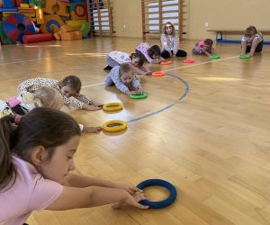 Dzieci w sali gimnastycznej ćwiczą z kolorowymi ringami, przesuwając je po podłodze.