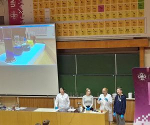Prowadzący i uczniowie na tle ekranu ukazującego pojemniki z kolorowymi płynami