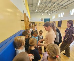 Starsza uczennica rozdaje słodycze młodszym dzieciom w sali gimnastycznej. Dzieci ustawione są w kolejce.