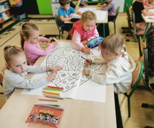 Kilka dziewczynek siedzi przy stoliku i wspólnie koloruje duży plakat z napisem „Każdy jest ważny”. Na stole leżą kredki i przybory szkolne.