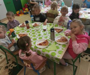 Grupa dzieci siedzi przy długim stole przykrytym kolorowym obrusem, na talerzach kawałki pizzy, obok stoją bidony z wodą.