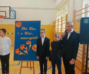 Czterech uczniów na tle tablicy dekoracyjnej, jeden z nich przemawia do mikrofonu