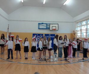 Uczniowie klasy czwartej i piątej przy mikrofonach, z małymi flagami Polski w dłoniach