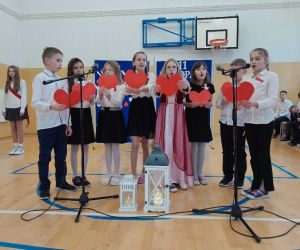 Uczniowie klasy czwartej przy mikrofonach, trzymają czerwone papierowe serca w dłoniach