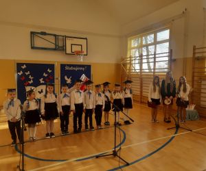 Uczniowie klasy pierwszej oraz uczennice klasy siódmej i piątej na tle tablicy dekoracyjnej