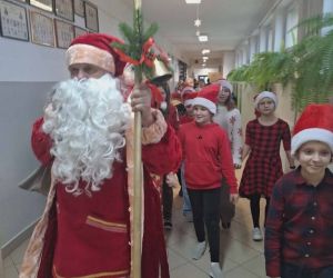 Mikołaj przechadza się szkolnym korytarzem, a za nim idą uczniowie w świątecznych czapkach.
