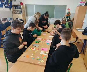 Uczniowie klas IV–VIII siedzą przy stolikach w sali lekcyjnej i wykonują kartki okolicznościowe dla Babci i Dziadka z kolorowego papieru.

