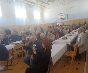 Babcie i Dziadkowie siedzący przy długich stołach na sali gimnastycznej, uczestniczący w uroczystości szkolnej. Na stołach przygotowany poczęstunek, a część gości obserwuje występy dzieci.