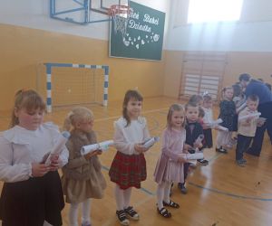 Grupa dzieci w eleganckich strojach stoi w rzędzie na sali gimnastycznej, trzymając w dłoniach zwinięte kartki. Dzieci przygotowują się do wręczenia upominków Babciom i Dziadkom.